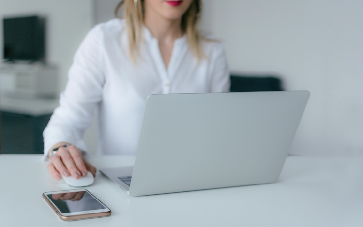 Eine Frau mit silbernem Laptop bedient eine Computermaus, daneben ein iPhone.