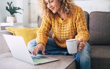 Junge Frau mit gelben Pullover sitzt am Sofa mit aufgeklappten Laptop auf dem Tisch.